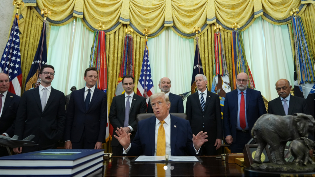 A photograph of Donald Trump speaking and gesturing with hands up from the Resolute Desk in the Oval Office. He is surrounded by a group of government officials and advisors. On his desk are a F-35 model and a Republican elephant statue. This image relates to Trump announcing the acceleration of national quantum technology innovation.
