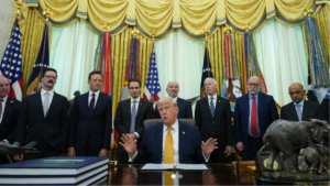 A photograph of Donald Trump speaking and gesturing with hands up from the Resolute Desk in the Oval Office. He is surrounded by a group of government officials and advisors. On his desk are a F-35 model and a Republican elephant statue. This image relates to Trump announcing the acceleration of national quantum technology innovation.